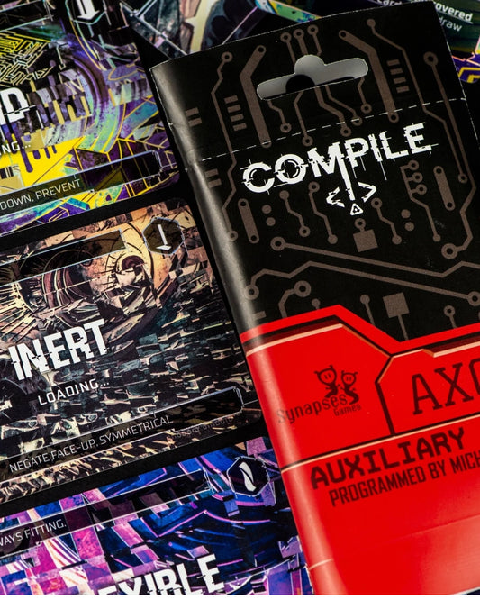 A close-up of a black and red Synapse Games 'Compile' auxiliary 3 product package, layered over several colourful printed cards or inserts with circuit-like graphics and text such as 'INERT' and 'AUXILIARY'.
