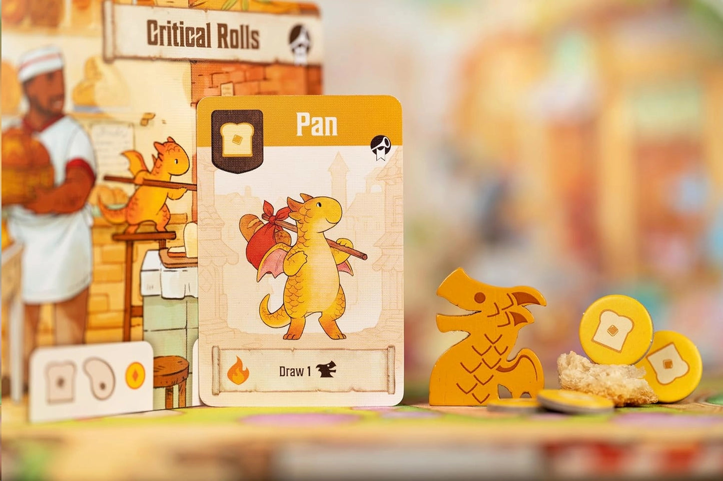 A tabletop close-up of a Flamecraft board game setup featuring a central card labelled 'Pan' with a cartoon dragon illustration, a yellow dragon token, gold coin tokens, and a blurred game board in the background with a baker character and 'Critical Rolls' text.