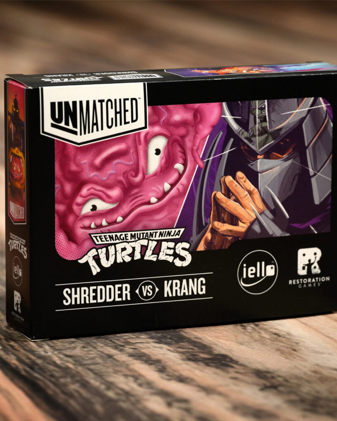 A boxed card game or collectible set titled "Teenage Mutant Ninja Turtles Shredder vs Krang" on a wooden surface. The box is black with large illustrated artwork of a pink turtle face and a dark armored figure, plus visible logos including Unmatched, iell, and Restoration Games.