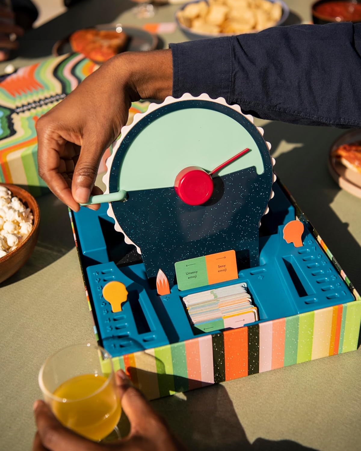 A colourful board game Wavelength by CMYK is being held by a person with snacks and drinks on a table