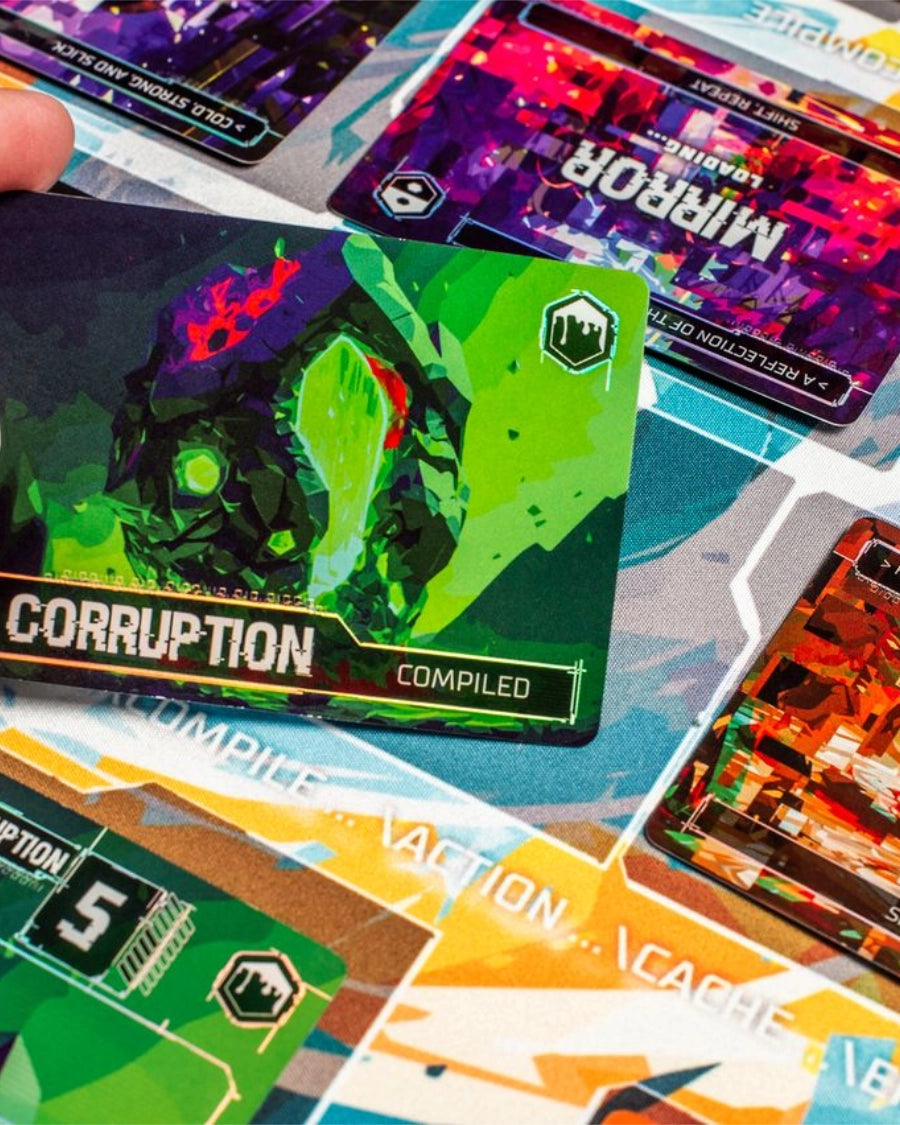 A close-up of Complie: Main 2 Card game board and cards, with a hand holding a green and black 'Corruption' card over a colourful board featuring other cards like 'Mirror' and 'Corruption'.