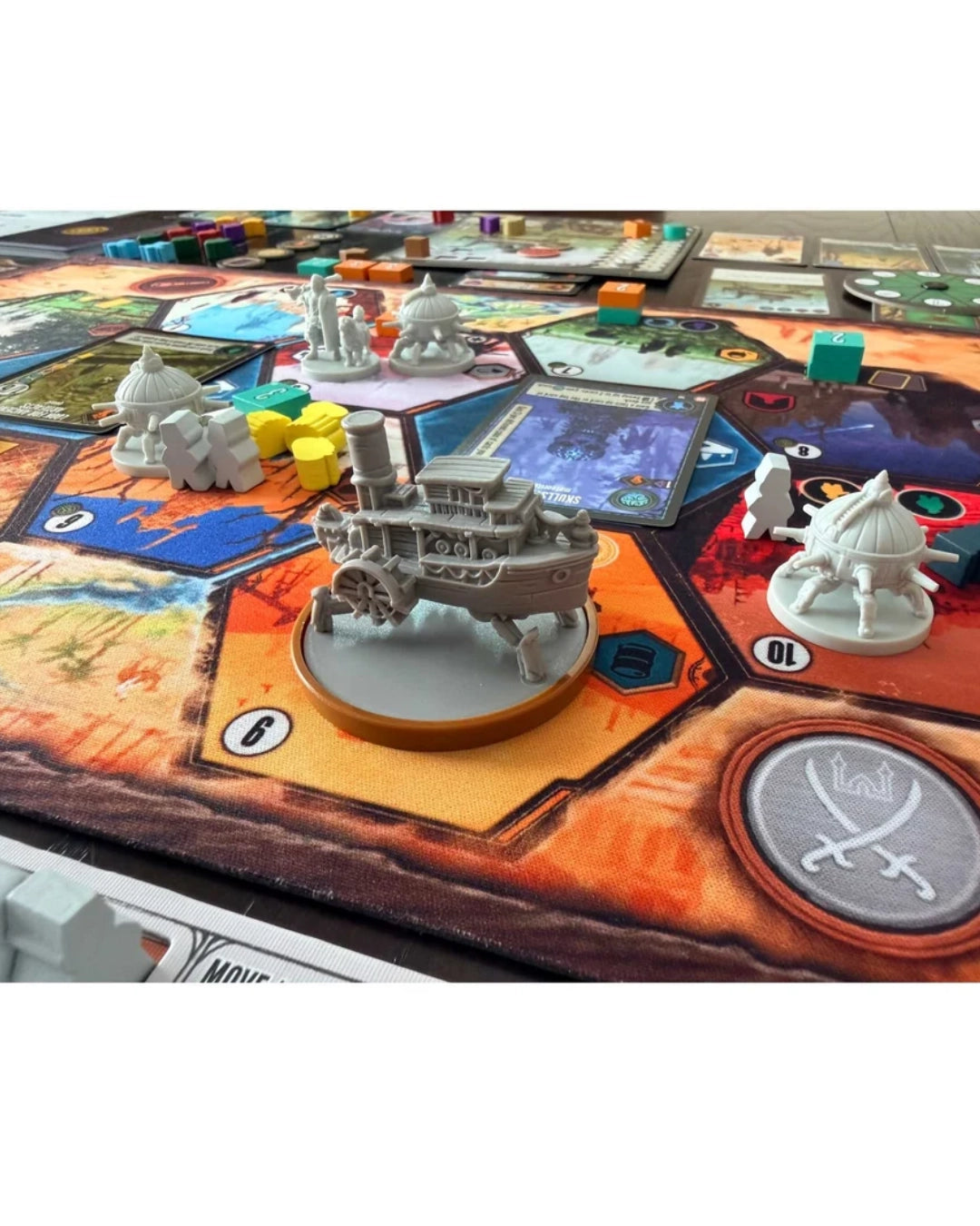 A tabletop gaming setup for Duel of Meloch: Scythe vs Expeditions showing a detailed grey steampunk-style miniature on a circular base, placed on a colorful fantasy board game mat with cards, tokens, and other small game pieces visible around it.