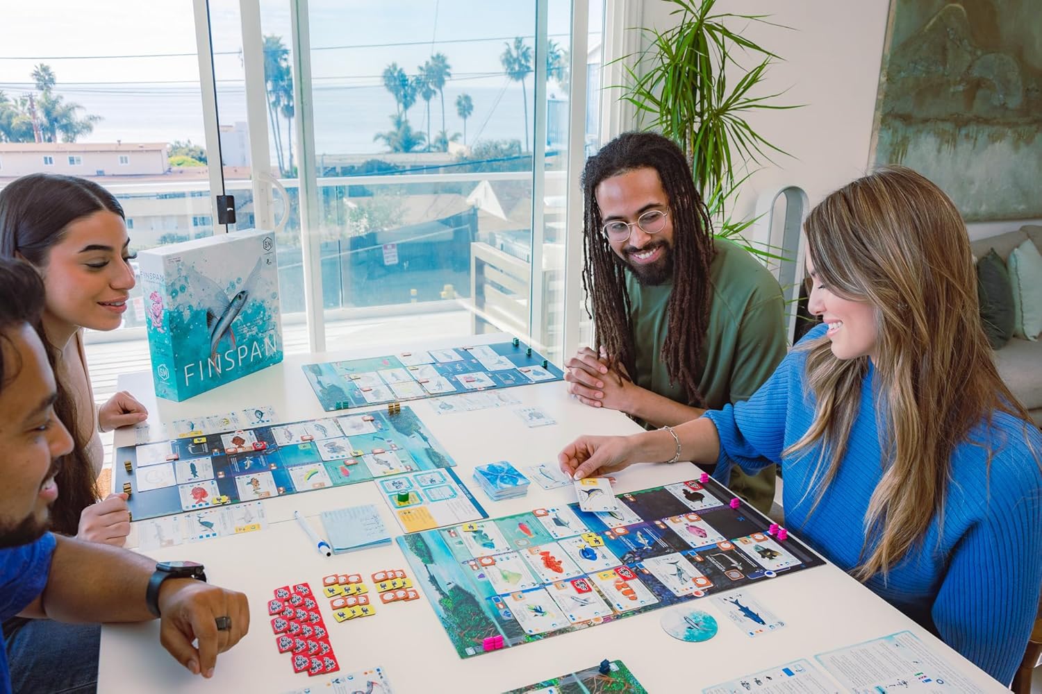 A group of people playing Finspan, the board game, together in a bright room with large windows.