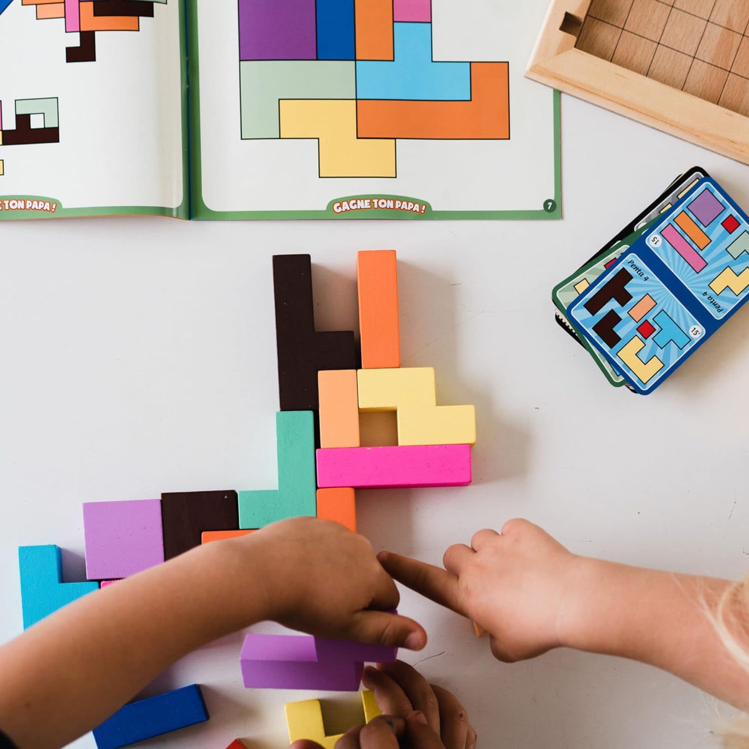 Two children are playing with colourful wooden blocks on a white surface, with a phone displaying a similar game in the background.