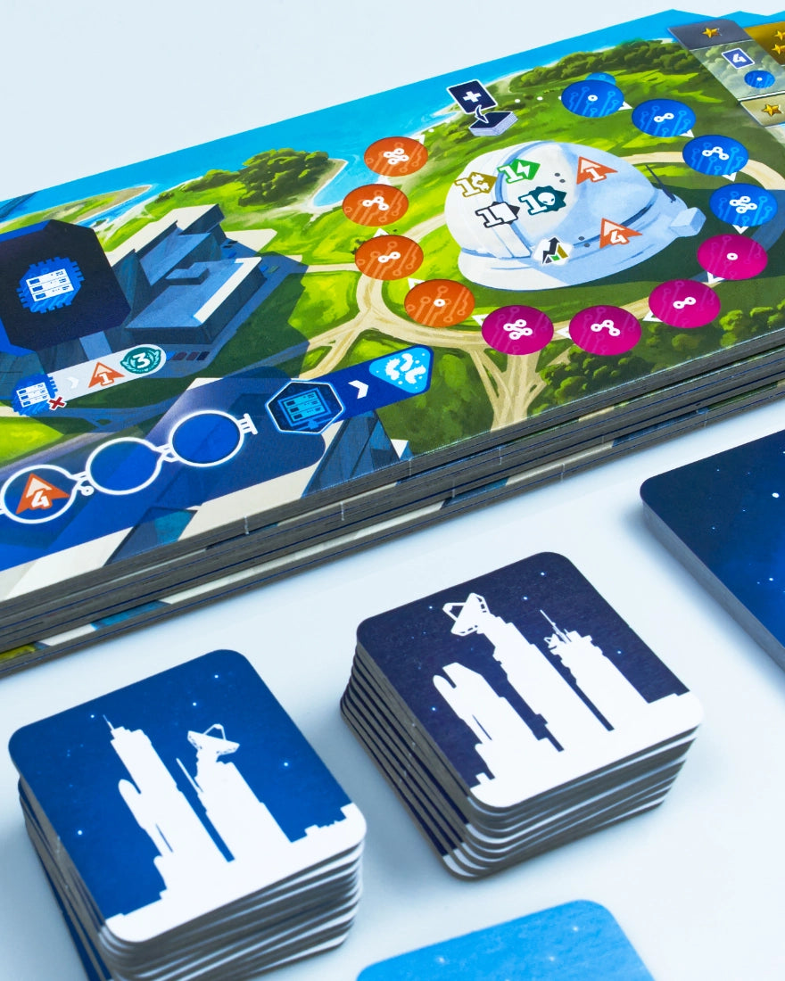 Close-up of SETI: search for extraterrestrial intelligence board game set with a colourful illustrated game board, blue and navy cards, and stacks of square cards featuring white building silhouettes on a light background.