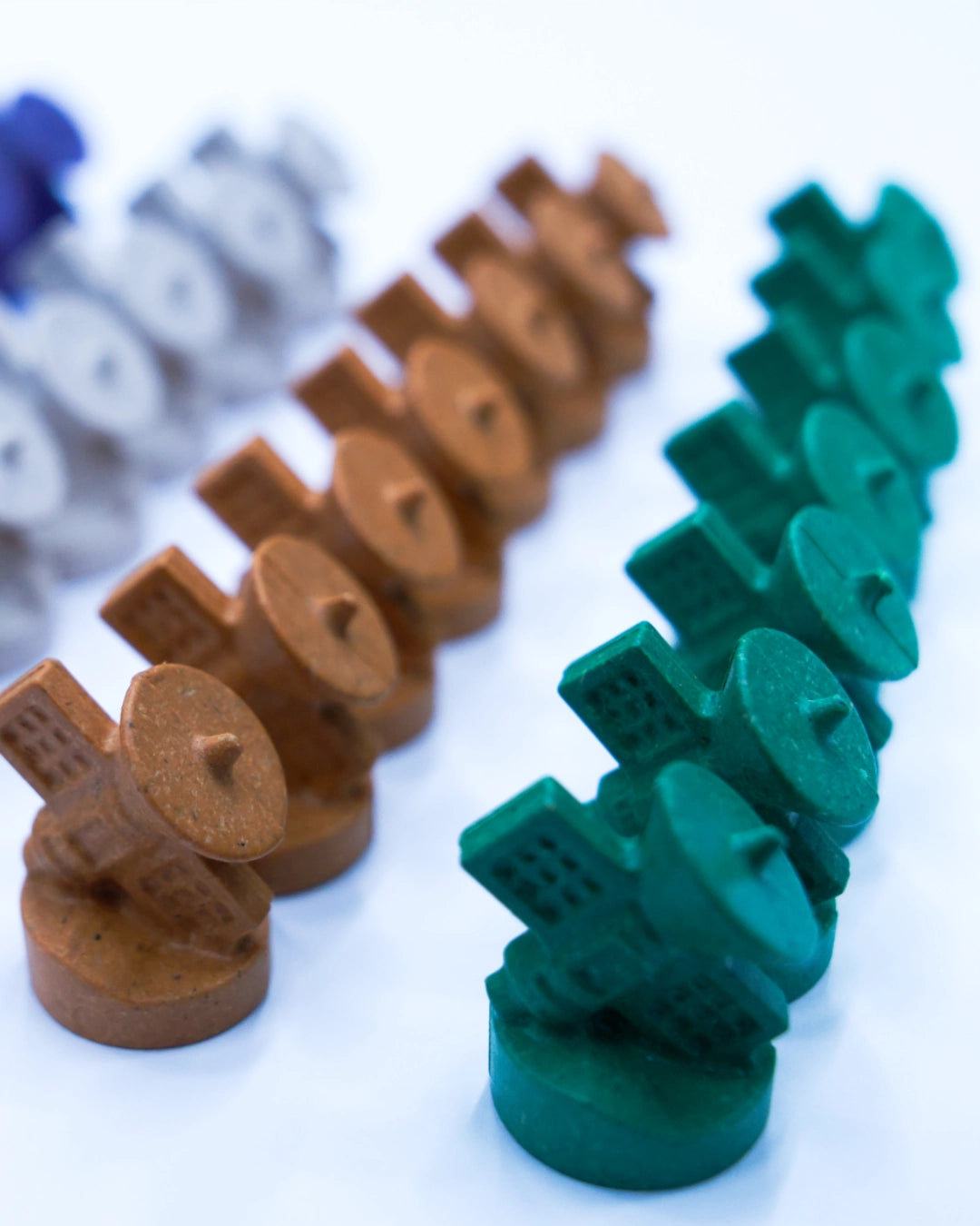 A close-up shot of small, round, castle-like toy pieces arranged in diagonal rows for SETI: search for extraterrestrial intelligence board game. The foreground pieces are a dark green colour, while the pieces behind them are a warm brown/tan, and a set of blue and grey pieces is blurred in the background on a white surface.
