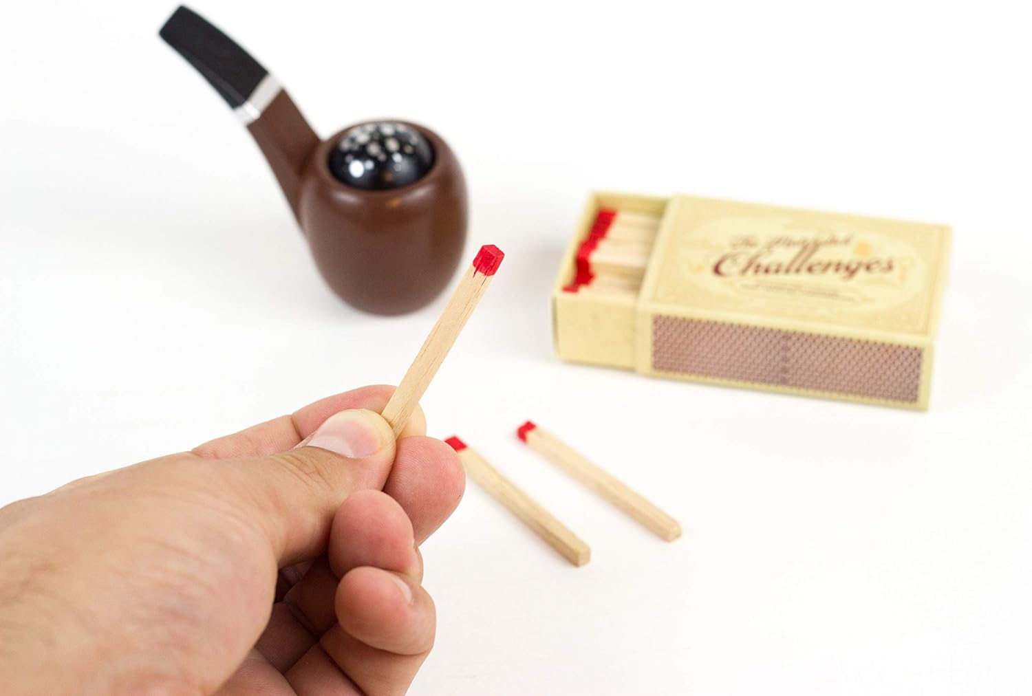 Hand holding matches next to a wooden matchbox and pipe on a white background
