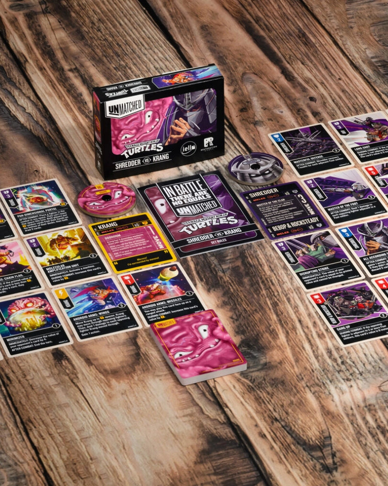 Unmatched Teenage Mutant Ninja Turtles card game components arranged on a rustic wooden tabletop, including the product box, a token, a die, and multiple cards spread out.