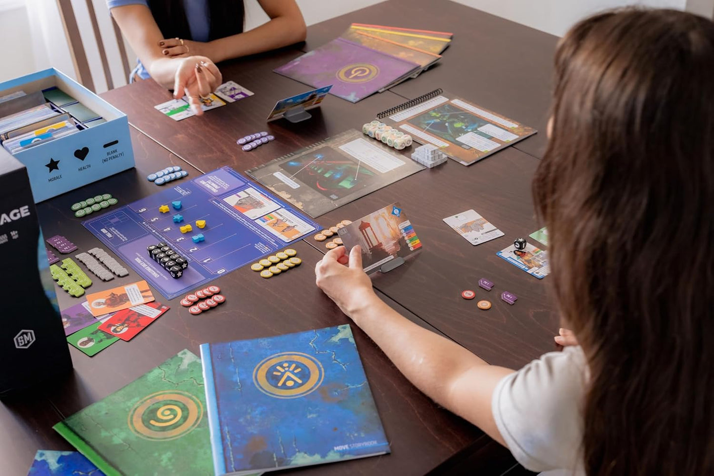 Two people playing a Vantage board game with various game pieces and cards on a table.