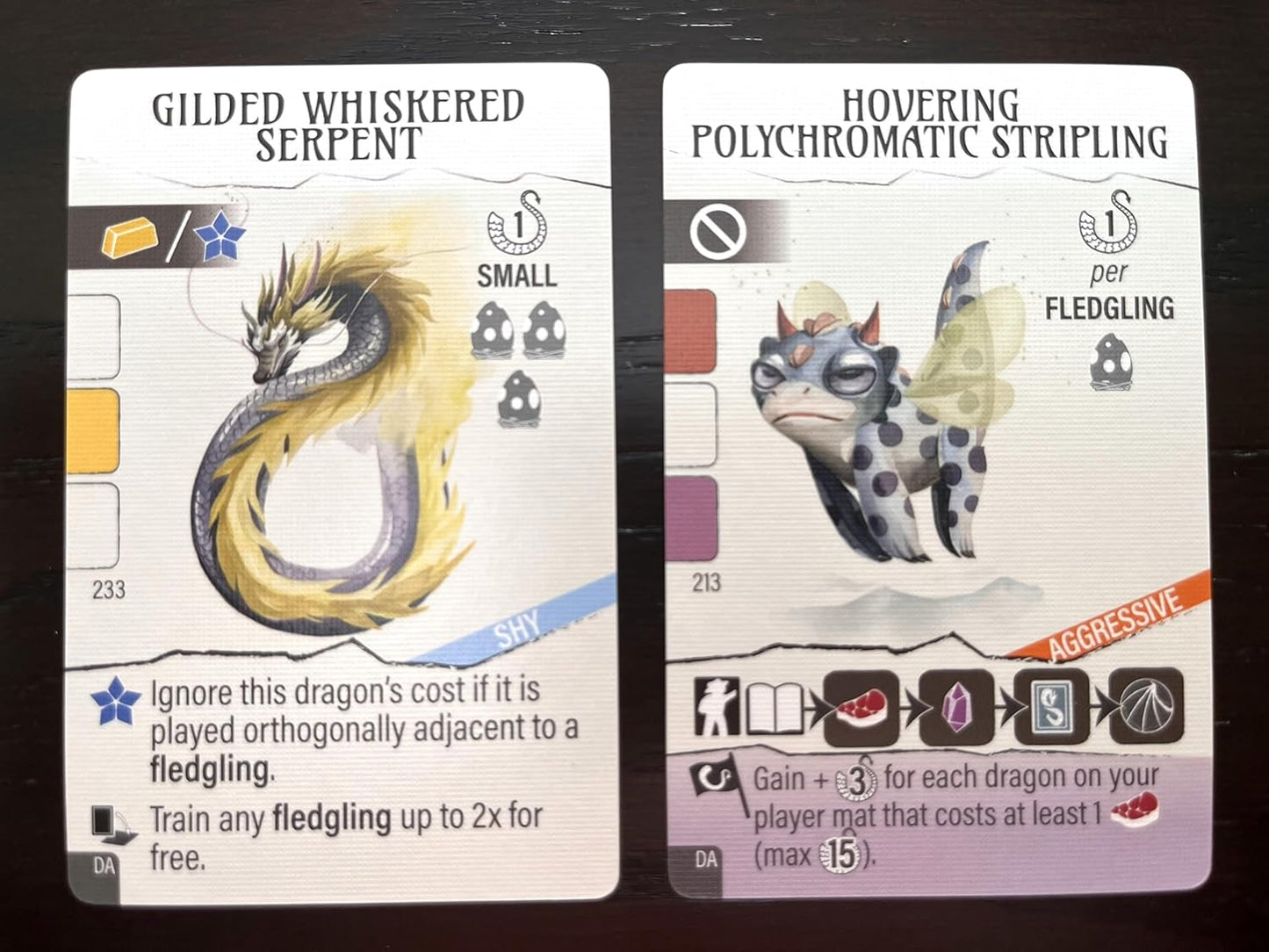 Two fantasy game cards featuring gilded whiskered serpent and hovering polychromatic stripling on a dark surface.
